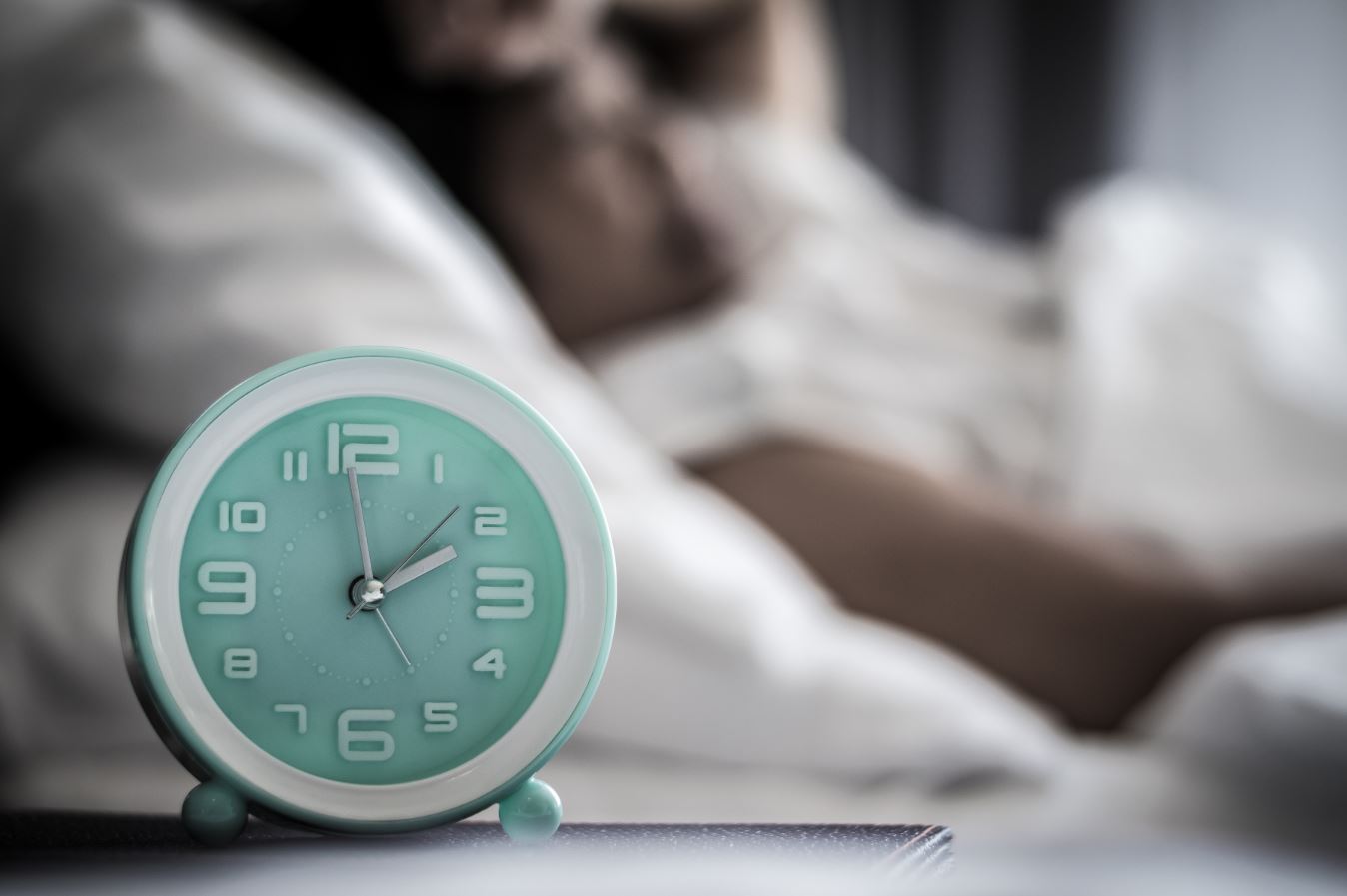 An analog clock on a nightstand shows the time at 2 o'clock. Behind it is a blurred-out bed and a person lying in it.