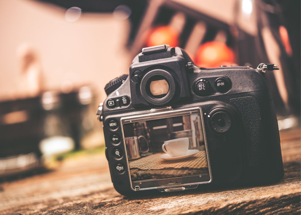 Displayed is a digital camera sitting on a table with a coffee cup being shown in the camera's preview display.