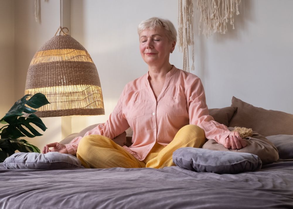 Displayed is an elder woman meditating in the lotus postion on a bed with soft cushions.