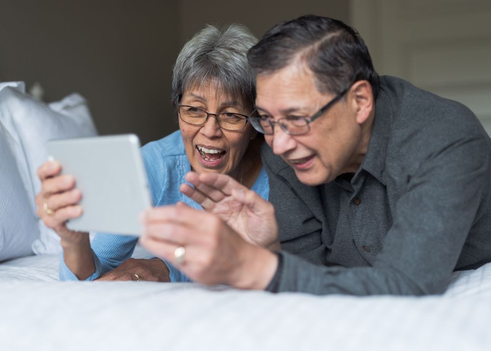 Displayed are two seniors lying on their bed and having a video chat on their tablet with loved ones.