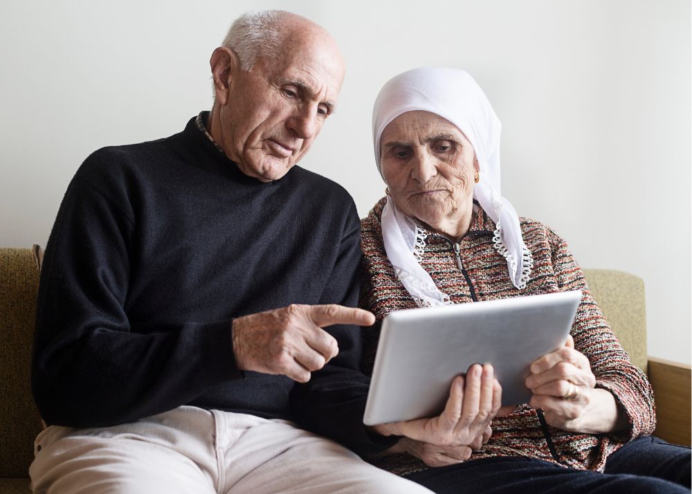 Displayed is an elder couple, with the woman wearing a head scarf, looking at a tablet together.